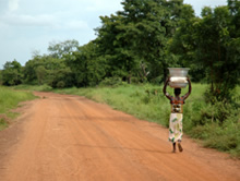 Woman carrying water bottles on her head along a long dusty road.